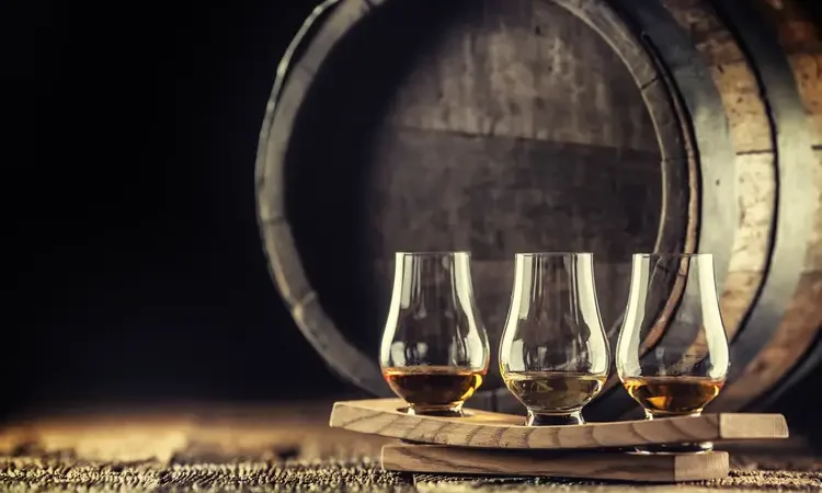 Glencairn whiskey tasting cups on a wooden serving, with a whisky barrel in the dark background